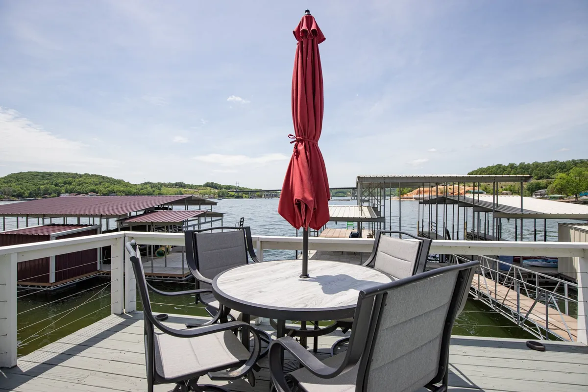 This image captures a table with an umbrella set up by the lake, with a dock area visible in the background. The setting suggests relaxed lakeside dining and outdoor socializing, with ample space for group activities and celebrations. The tranquil water and open sky create an inviting atmosphere.
