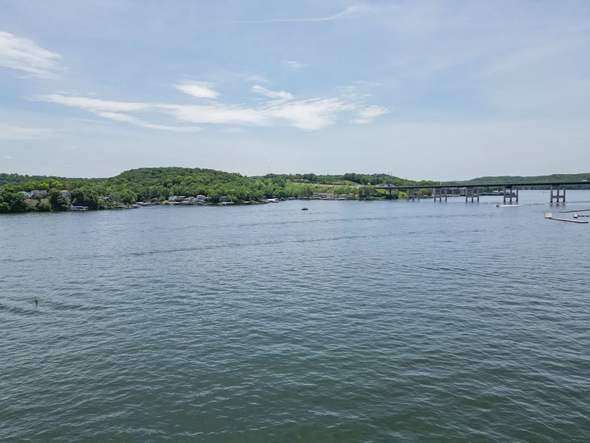 A sweeping view of calm lake waters surrounded by lush green hills, with a distant bridge and small boats dotting the scene. The landscape is bathed in peaceful, natural light, capturing the serenity and recreational opportunities of the Lake of the Ozarks area.