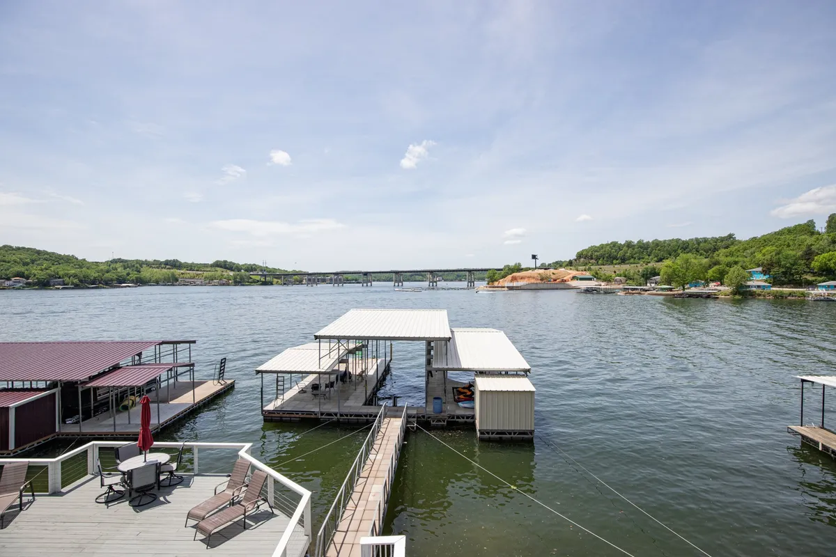A tranquil lakeside setting featuring a dock, boathouses, and lush surrounding greenery. The scene is set for boating and fishing, while the expansive water and sunset lighting evoke relaxation and social gatherings by the shore.