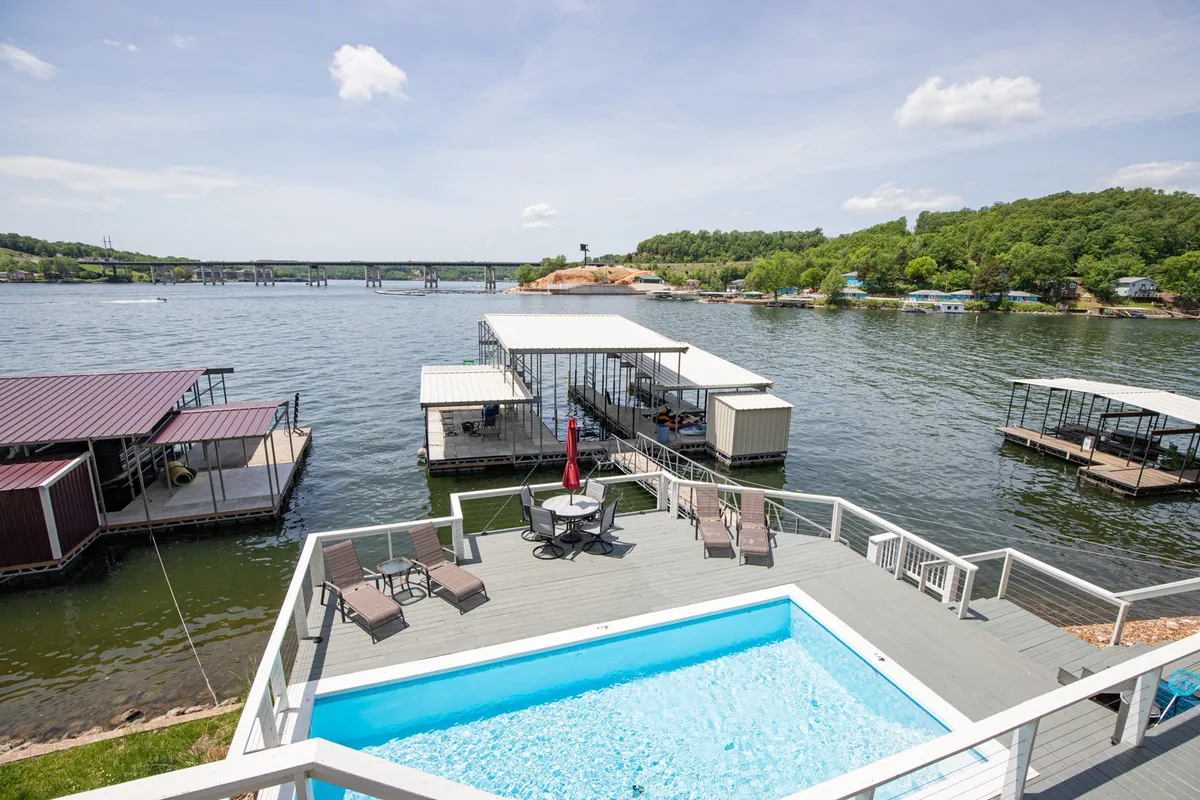 The hero image showcases a sparkling private saltwater pool overlooking the serene lake, with a spacious outdoor dining set ready for meals al fresco. Beyond the pool, an expansive dock with boats invites guests to enjoy the water, all framed by lush green hills and the promise of breathtaking sunset views.