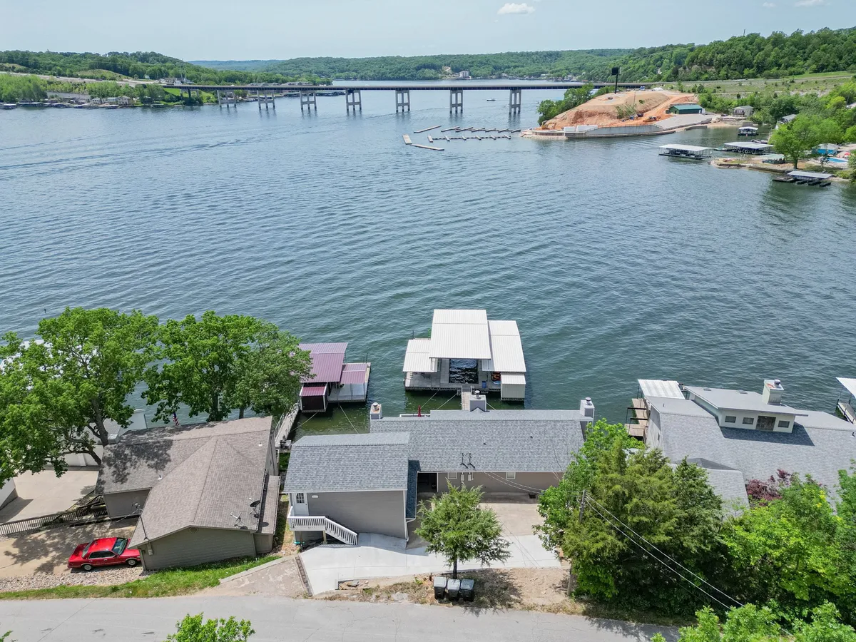 A scenic lake view capturing calm water, docked boats, a bridge in the background, green trees along the shore, and houses lining the waterfront. The image evokes the inviting atmosphere of morning fishing, water sports, and family gatherings by the water.