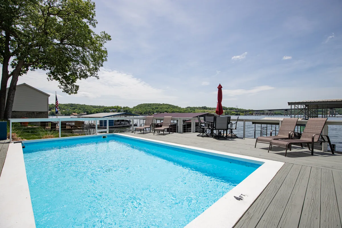 A sparkling swimming pool surrounded by lounge chairs and a striking red umbrella sits on a deck overlooking the lake. The vibrant blue water invites guests to swim and play, while the serene lake view in the background creates a picturesque resort-like atmosphere. The poolside area appears perfect for both sunbathing and lively family activities.