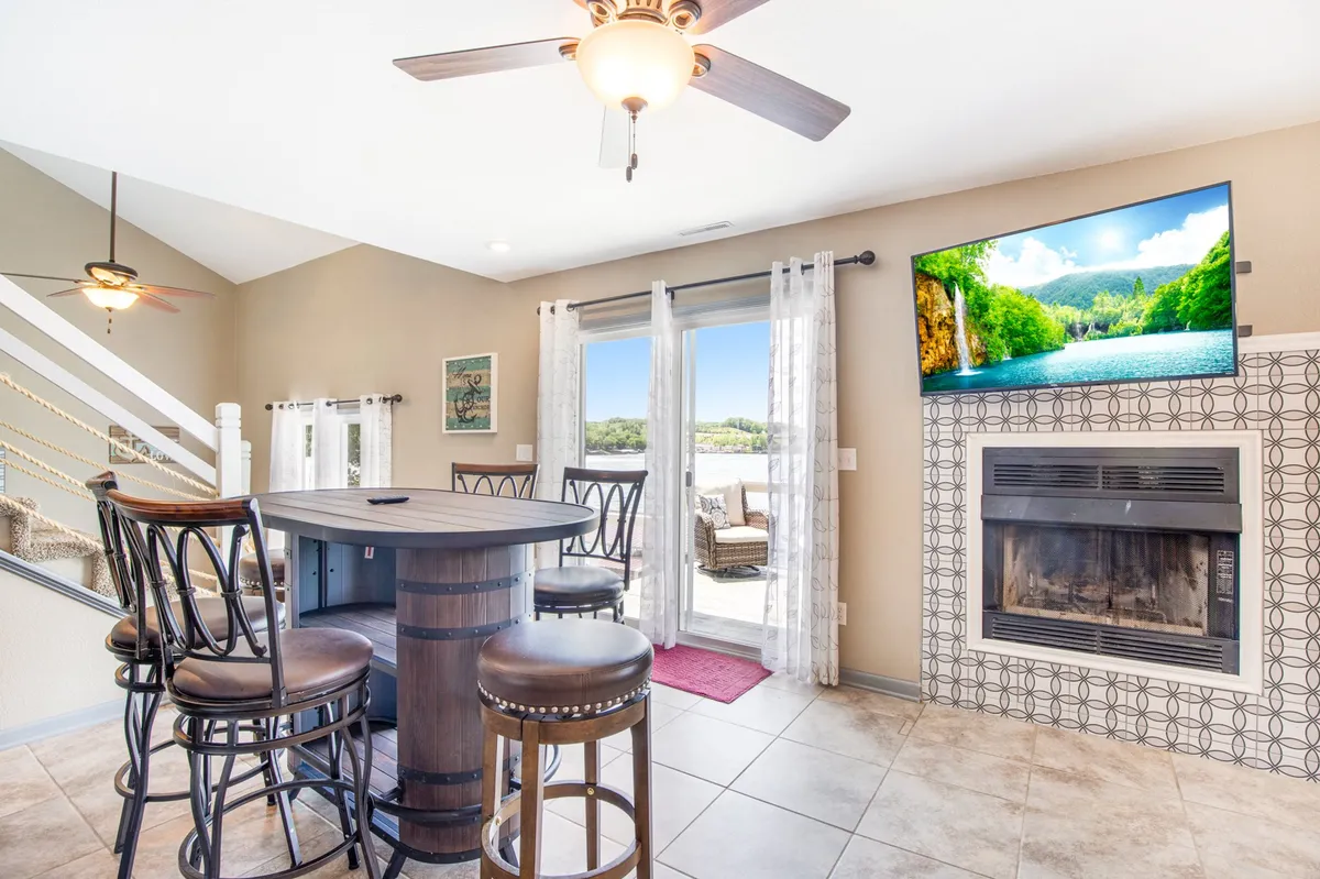 A cozy interior dining and lounging space featuring a round dining table, barstools, a fireplace, large windows, and a television. The area is bathed in natural light, with ample seating for socializing or sharing a meal. The view through the windows hints at the scenic outdoors and brings in abundant daylight.