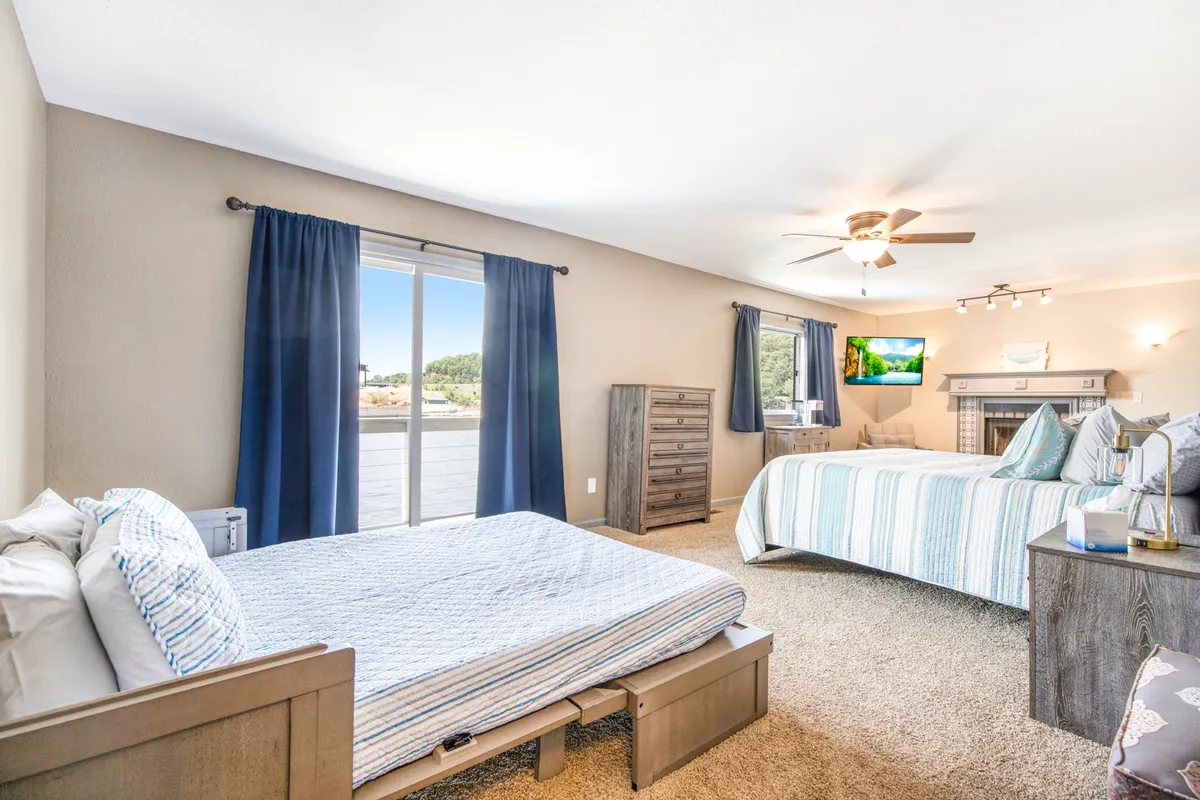 A bright bedroom with two beds dressed in striped bedding, a large window with blue curtains offering glimpses of the outdoors, a wall-mounted TV, and a wooden dresser. The setup is perfect for guests or families, allowing for socializing, relaxation, and enjoying lake views from bed.