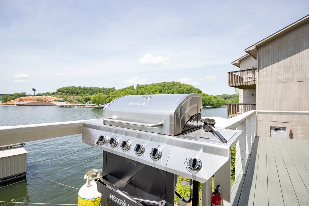 A well-equipped outdoor grill set on a deck with a scenic water view and green hills in the distance. The inviting setup suggests summertime barbecues and al fresco dining, capturing the essence of outdoor entertainment by the lake.