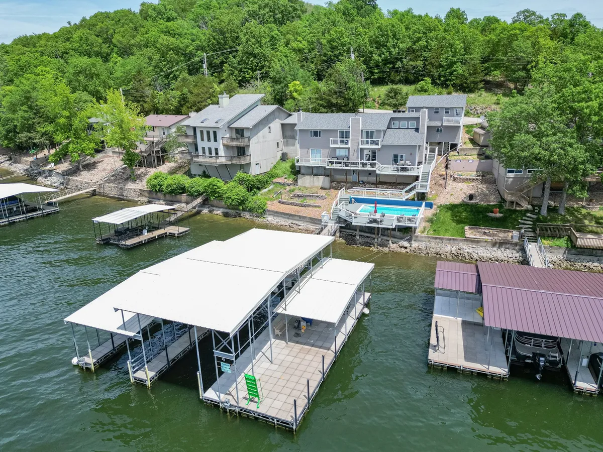 A wide angle exterior shot featuring several houses along the lake shore, a swimming pool, boathouses, multiple docks, and lush greenery. The scene captures the inviting dock and the vibrant lakefront, with ample space for sunbathing, swimming, and launching watercraft.