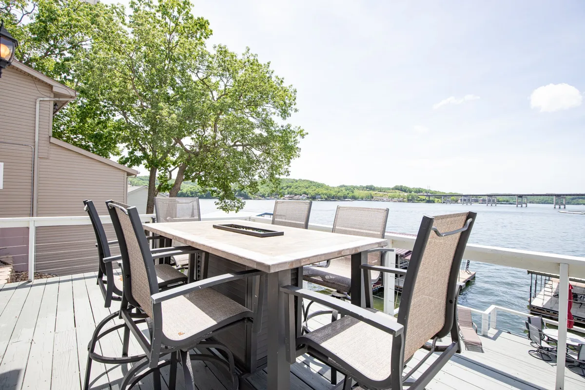 An outdoor dining area set against a beautiful lake view, surrounded by greenery. The table is ready for family or group gatherings, with the outdoors seamlessly connected to the interior spaces through nearby windows and doors.