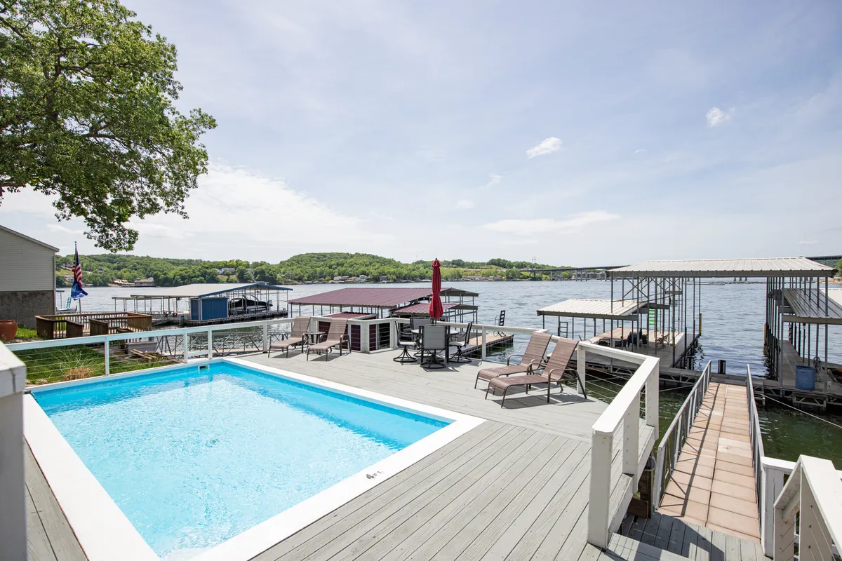 A wide-angle exterior shot capturing a sun-soaked patio with ample outdoor seating, a sparkling resort-style swimming pool, and an expansive view of the lake. The dock and lounge areas are visible in the background, connecting the outdoor living spaces with the water.