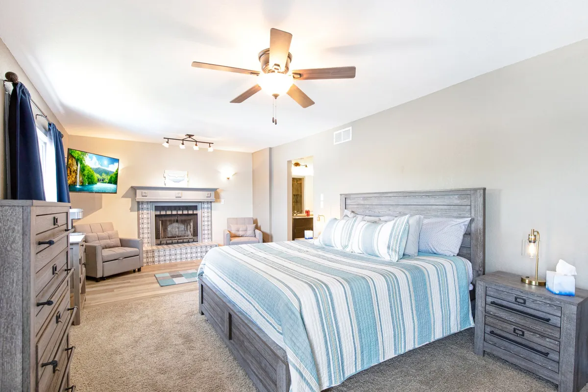A serene bedroom featuring a king-sized bed with striped bedding, a display-only fireplace, wall-mounted TV, and armchairs set against soft lighting. The arrangement exudes luxury and provides options for quiet relaxation, movie nights, or reading by the gentle glow of the fireplace feature.