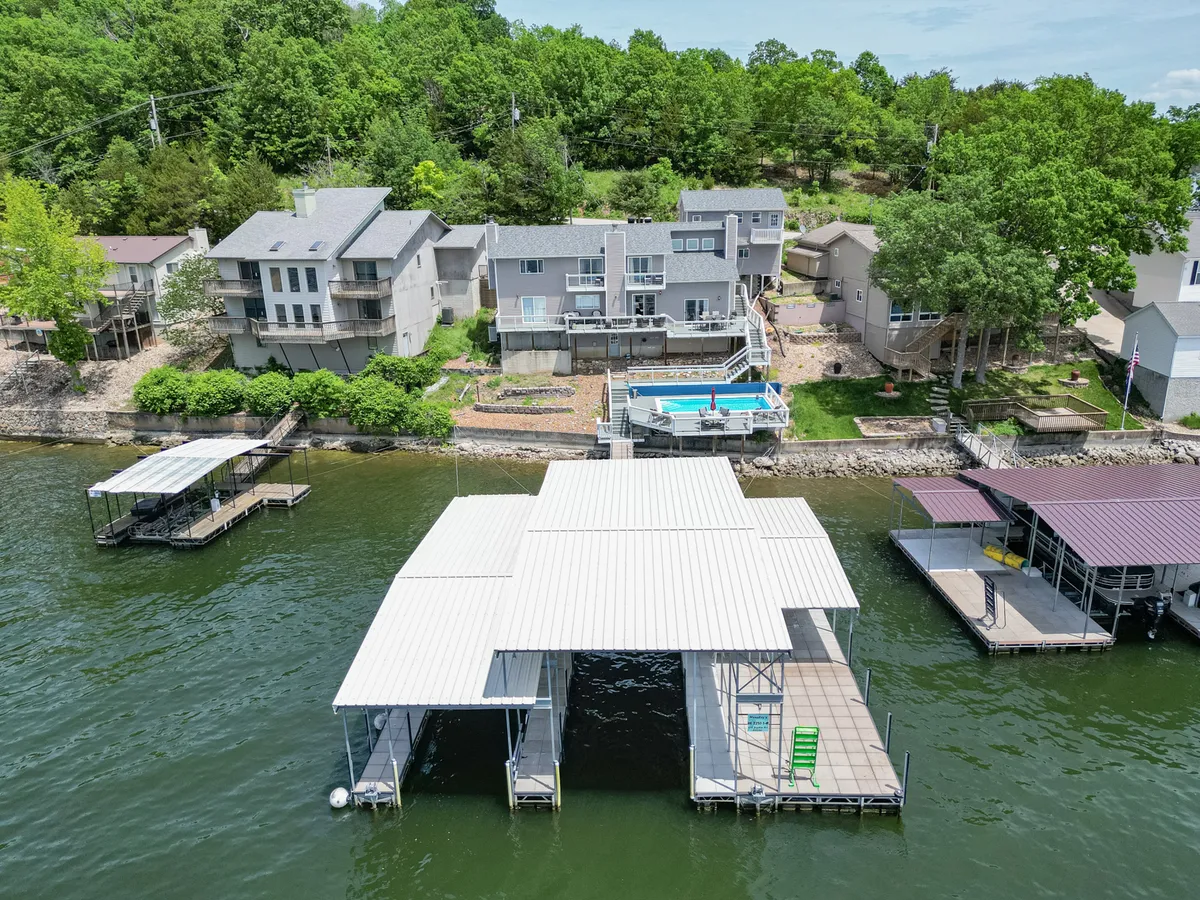 An exterior perspective showing a large dock extending into the lake, an adjacent swimming pool, wooded areas, and surrounding homes. The image highlights the spacious dock, calm waters, and areas for both boating and swimming.